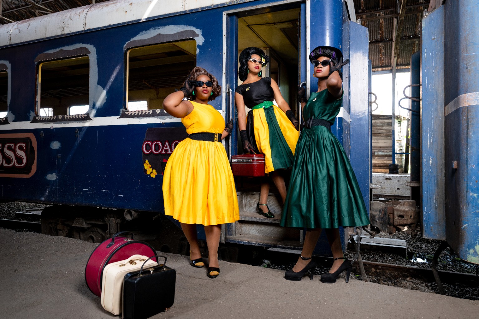 Three women dressed in vintage-inspired outfits with luggage beside a train